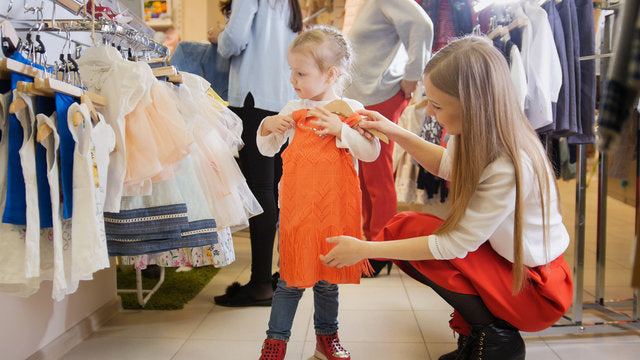Child holding a Cocoknut orange perfet fit dress in a kids wear store with clothing racks in the background.